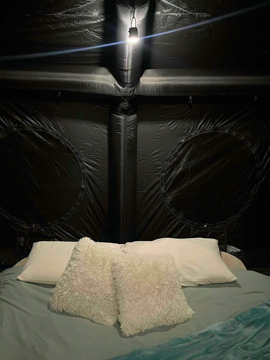 Interior of the blacked-out inflatable tent showing two round wall vents, a hanging LED lamp and a bed made up with pillows