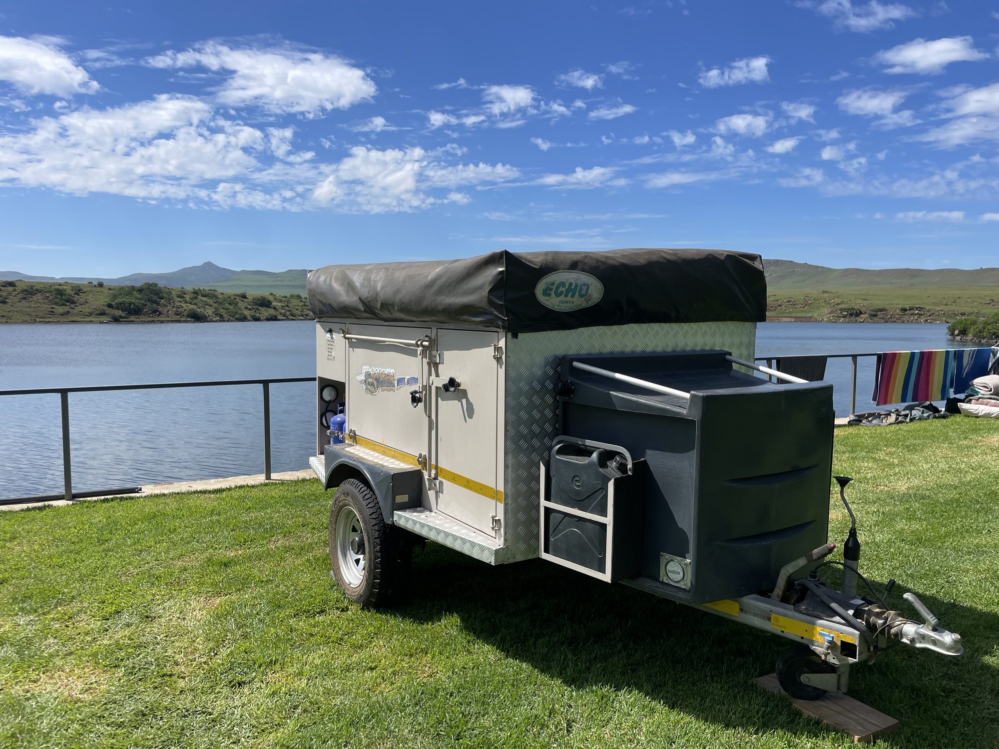 Off-road camping trailer with jerry cans and towels drying on a line beside a Drakensberg dam