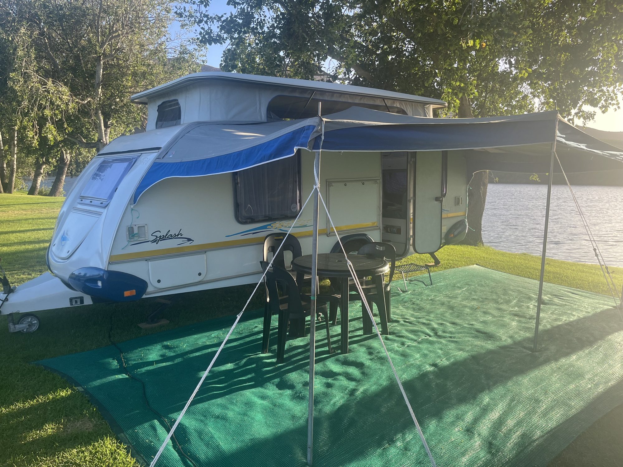 Caravan set up with awning, camp chairs, a table and a ground mat next to a dam at golden hour in South Africa