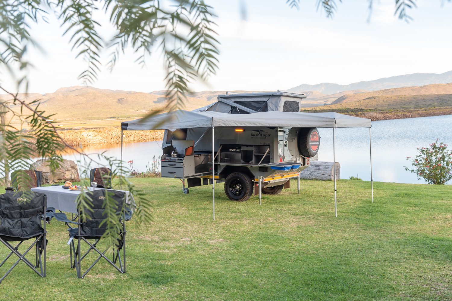 Off-road caravan with the awning out, camp chairs and a set table beside a calm dam