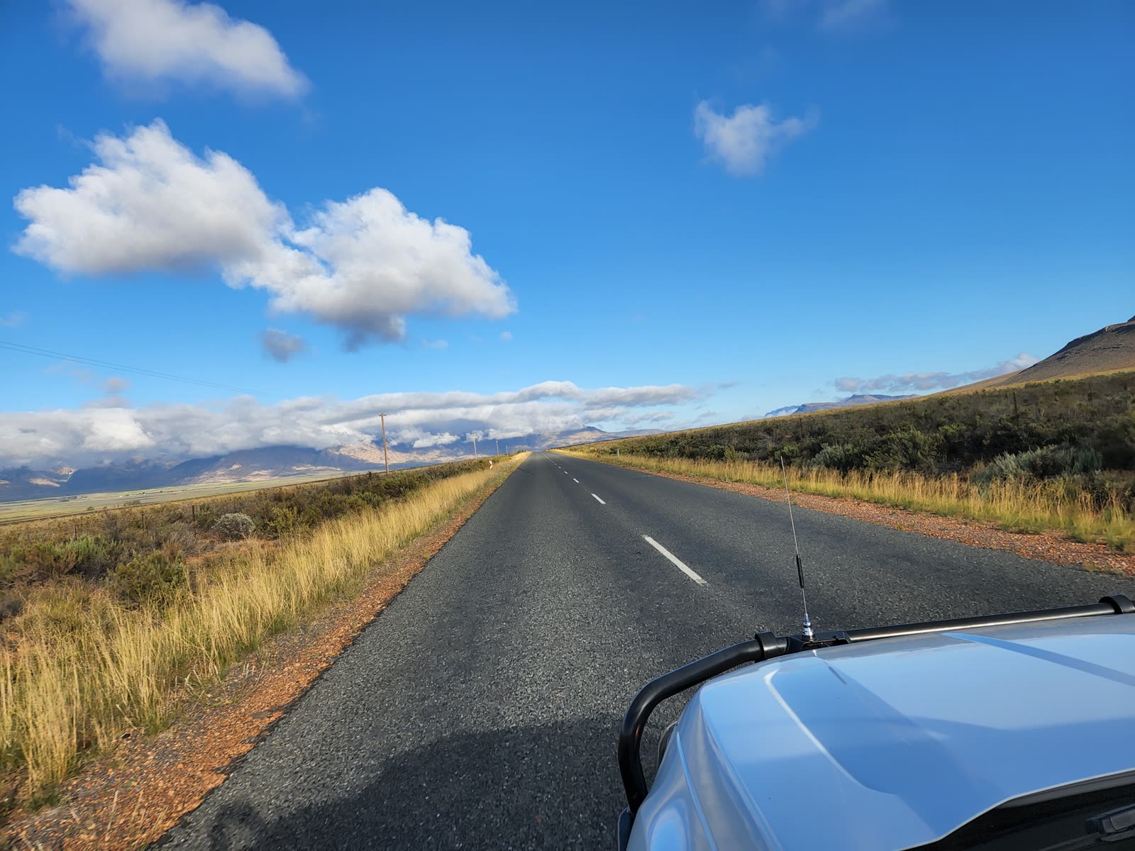 An empty tar road running towards Cape mountains under a blue sky with scattered cloud, seen over the bonnet of a tow vehicle