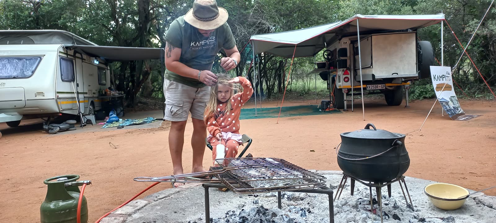 A father and small daughter at a bushveld campfire with a potjie on the coals, a caravan on one side and an off-road trailer with its awning out on the other