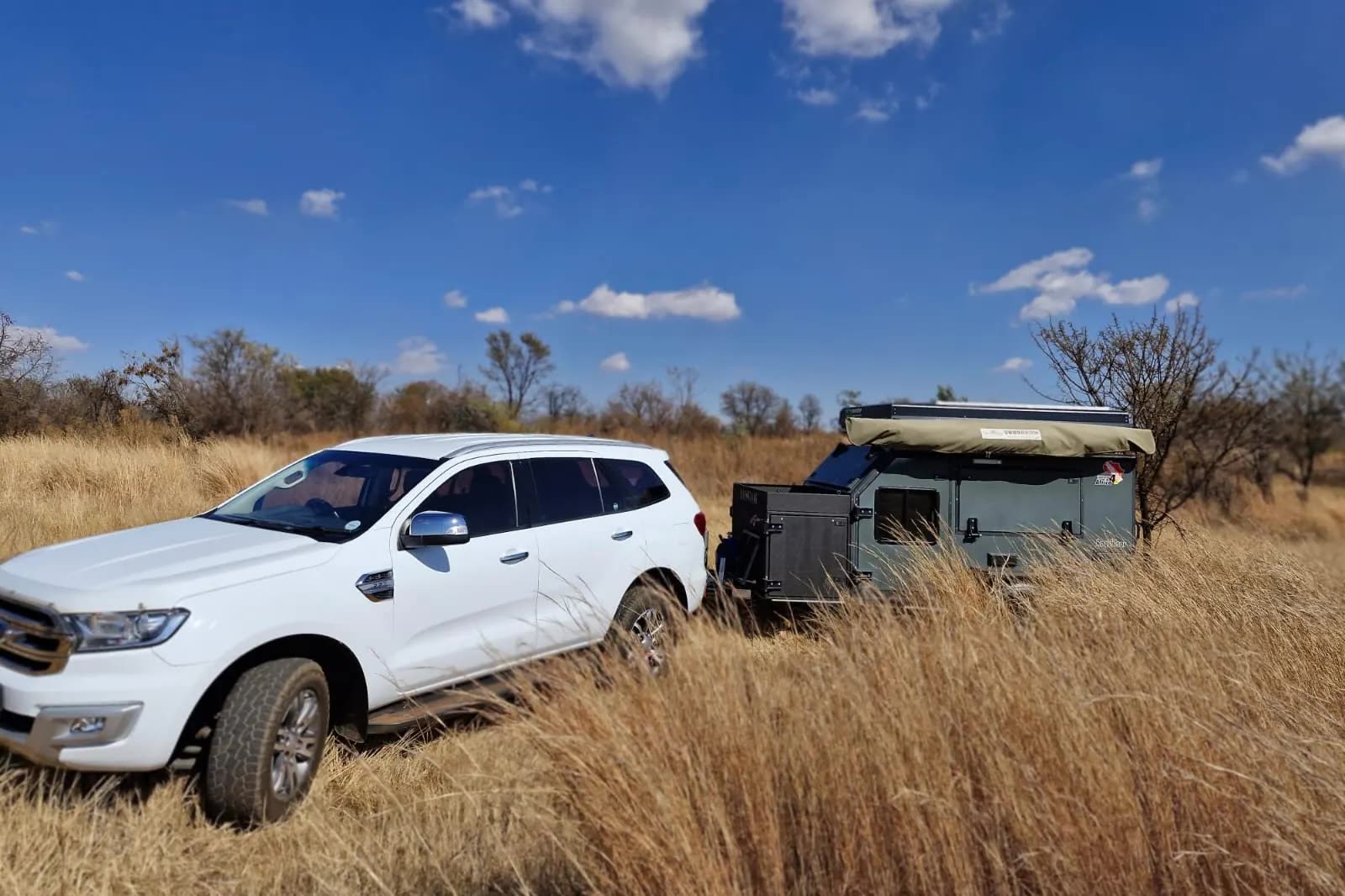 A white Ford Everest towing an off-road caravan through long dry grass in the bushveld