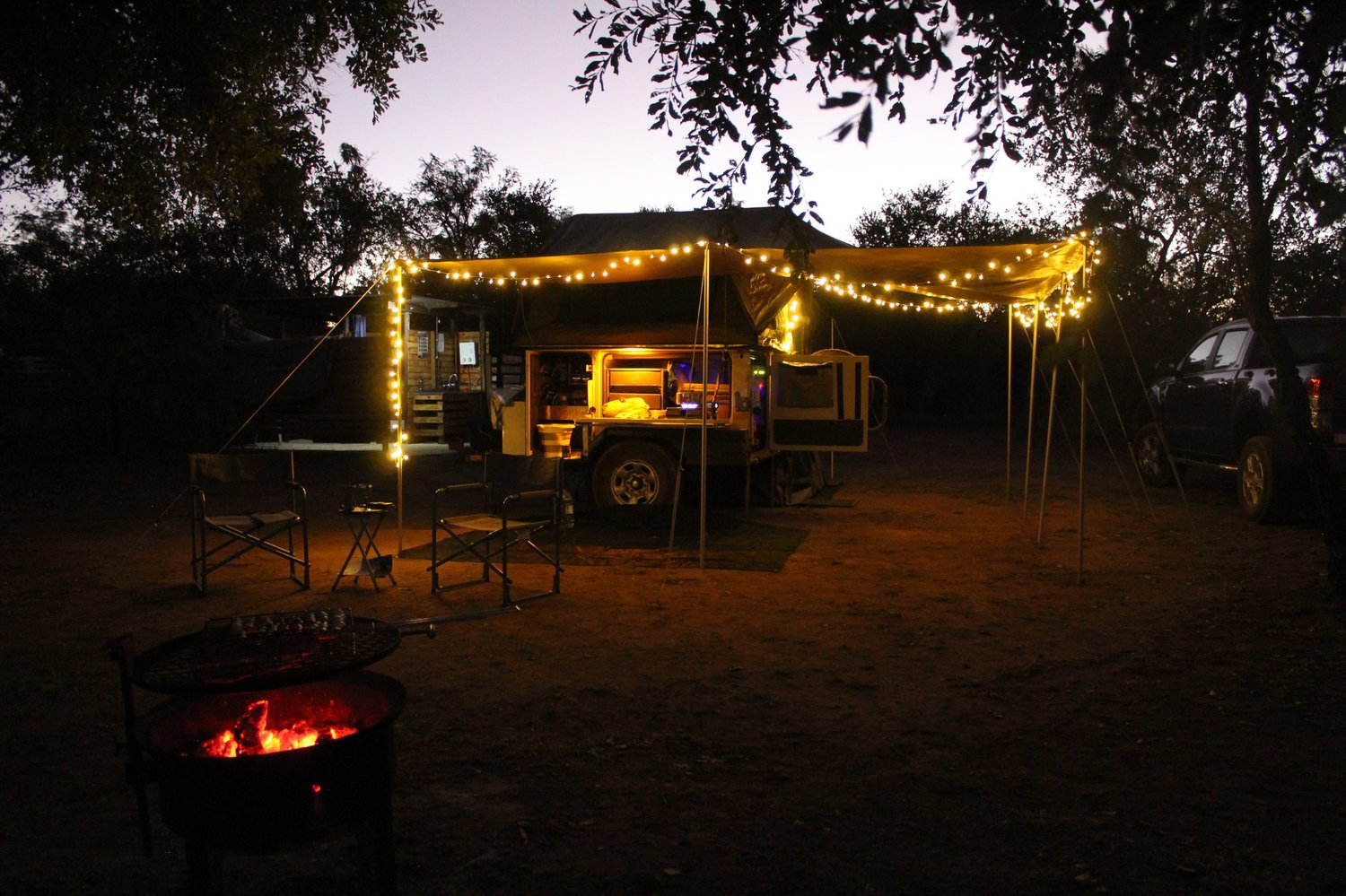 Camp lit up at night with a fire in the foreground, fairy lights and a glowing trailer kitchen