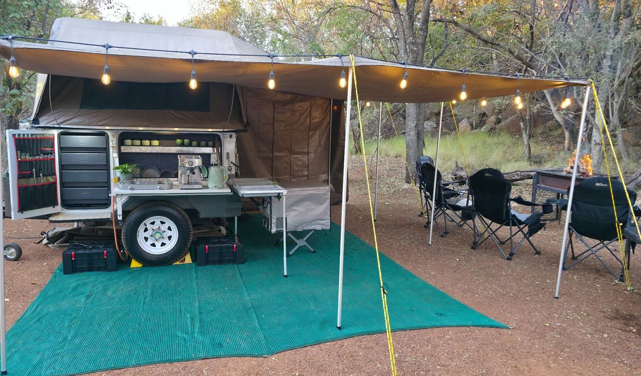 Camp set up under a tree at dusk with awning, string lights, camping chairs and a green groundsheet